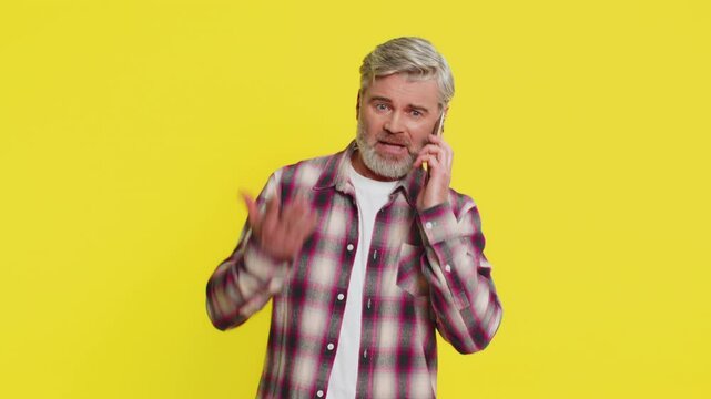 Mature man speaks on mobile phone with forceful tone, revealing anger agitation during tense talk quarrel. Middle-aged guy on yellow background shows argumentative stance, emphasizing heated dispute.