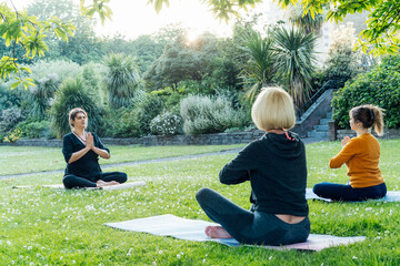 Female yoga class in park. Group of diverse women doing stretching pose exercising together with instructor on green grass lawn. Sport activity for health, wellbeing, mental health. Selective focus