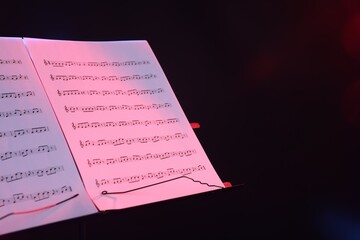 Stand with music sheets in neon lights on dark background, closeup. Space for text