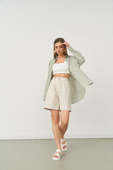 Stylish woman in casual outfit, wearing a white crop top, beige shorts, and an open green shirt. Her hand touches her hair, sandals complete her relaxed look, posing in studio