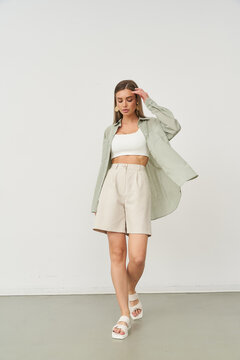 Stylish woman in casual outfit, wearing a white crop top, beige shorts, and an open green shirt. Her hand touches her hair, sandals complete her relaxed look, posing in studio