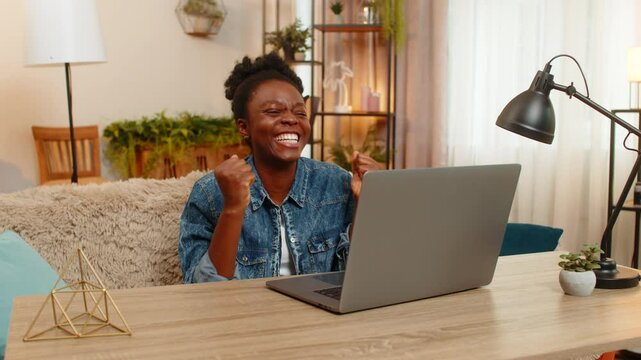 African American young woman celebrates good results from an online test and victory in a digital laptop game while sitting at table on home sofa. Black girl joyfully reacts to success, uplifting news