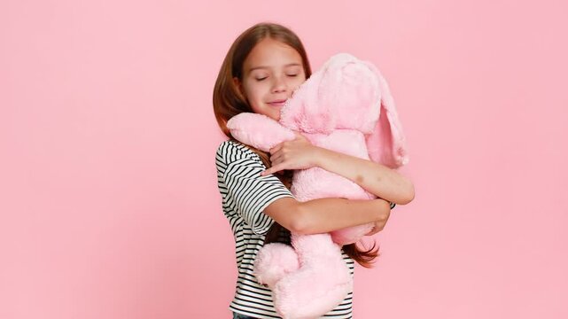 Young child girl gently hugs a plush toy bunny with love and happiness, smiling sweetly and closing eyes in joy, expressing warmth and emotional connection. School kid isolated on pink background.