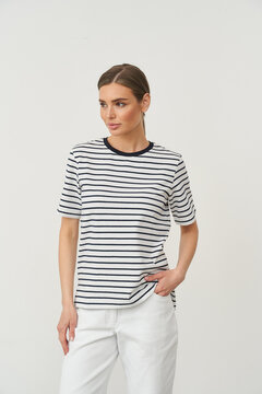 Striped short-sleeve shirt with a round collar worn by a person having light brown hair, styled in a neat side part. Person stands casually with one hand in white pants pocket