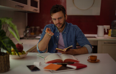 Young man in glasses gestures while holding a notebook, sitting at a kitchen table with open books,...