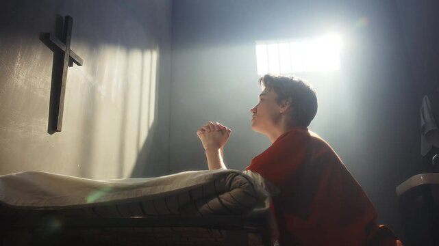 Female prisoner in orange uniform kneeling beside bed with hands clasped in prayer, looking at wooden cross on wall and praying under beam of sunlight from barred window