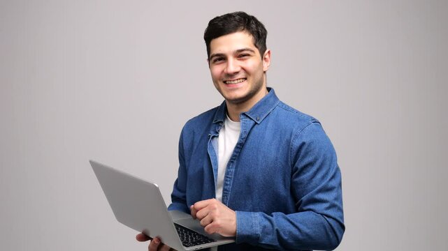 A cheerful young man engages with a laptop, embodying a spirit of innovation and approachability in a simple backdrop.
