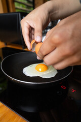 Close-up of hands holding cracked eggshell above a frying egg in a black pan. The yolk and white cook gently on the stove, capturing a simple breakfast preparation.

