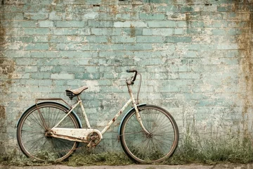 Fototapete A vintage white bicycle leans against a weathered, peeling blue brick wall with overgrown grass at its base. © Tina