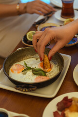 A close-up of a woman's hand dipping bread into fried eggs on a wooden table set for breakfast at a hotel. Cozy and appetizing morning moment
