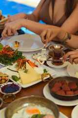 Close-up of a woman's hands cutting cheese with knife and fork at a hotel breakfast table set with salad, tea, and cheese. Pool visible in the background