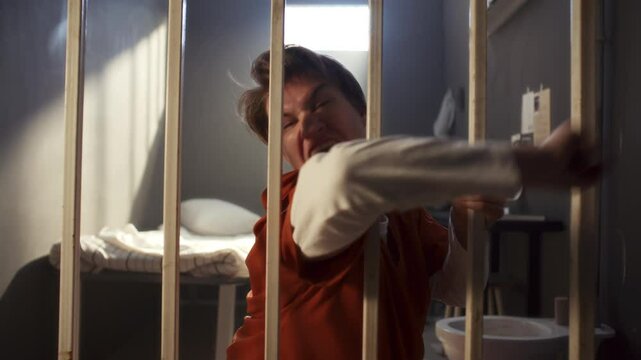 Furious female inmate in orange uniform running towards camera, screaming in rage and swinging arm through bars in burst of anger in prison cell