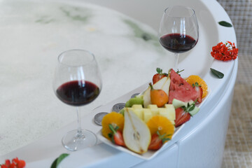 Elegant hotel jacuzzi filled with foam and roses, with wine glasses and fresh fruits on the edge