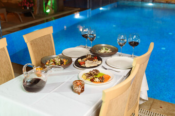 Elegant evening meal by the pool featuring wine glasses, grilled meat with vegetables, and ambient evening lighting
