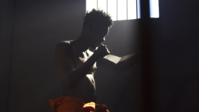 Shirtless Black male inmate practicing shadowboxing during workout in prison cell with light streaming through barred window