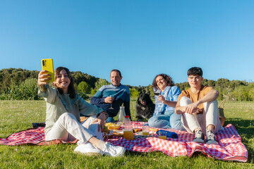 Family enjoying picnic in park with dog, taking selfie with smartphone on sunny day