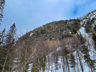  Rocky mountainside with patches of snow and ice, framed by bare trees and evergreen pines. The...