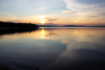 Obraz premium Stunning sunset over a calm lake with golden reflections, mountains on the horizon, and forest silhouette. Perfect for travel, nature backgrounds, peaceful landscapes, eco tourism, and inspiration.