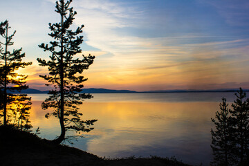  Beautiful lake landscape at sunset with calm water, silhouetted pine trees, colorful sky, and...