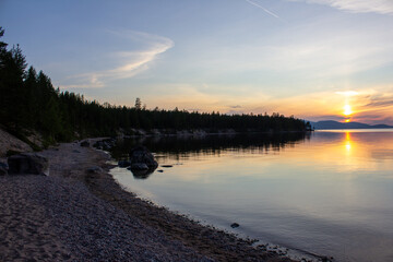  Beautiful sunset over tranquil lake with rocky beach, forest, and clear sky reflecting in still water. Serene nature landscape perfect for backgrounds, travel, relaxation, banners, and design project