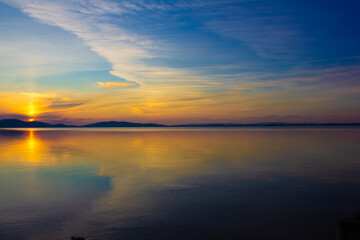 Obraz premium Stunning sunset over calm lake with vibrant colorful sky, dramatic clouds, and perfect reflection in water. Serene nature landscape ideal for backgrounds, banners, travel, meditation, and relaxation.
