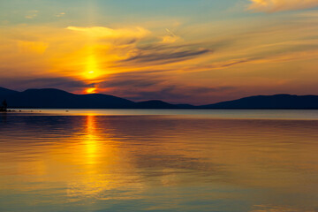Obraz premium Golden sunset over tranquil lake with colorful sky and smooth reflections. Serene evening scene with mountain silhouettes, glowing horizon and peaceful nature. Perfect for travel and landscape themes.