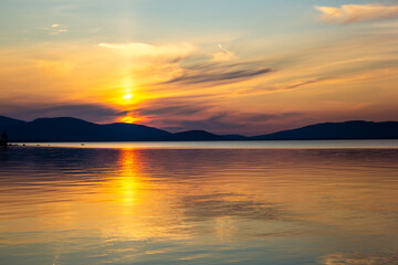 Obraz premium Golden sunset over tranquil lake with colorful sky and smooth reflections. Serene evening scene with mountain silhouettes, glowing horizon and peaceful nature. Perfect for travel and landscape themes.