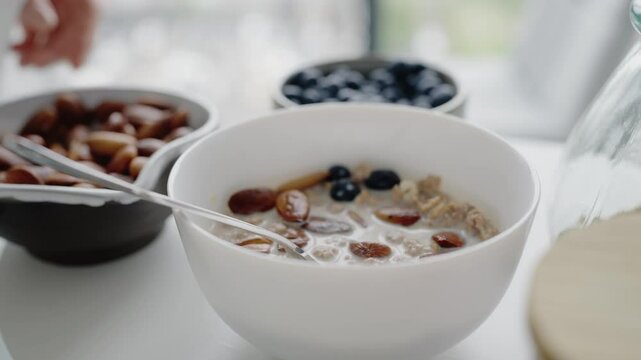 A hand adding nuts and berries to a white bowl of nutritious breakfast cereal, promoting healthy eating habits and a wholesome lifestyle. Hand Adding Nuts And Berries To Healthy Breakfast Cereal