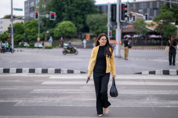 Young businesswoman walking on a crosswalk in the city, holding a smartphone and a bag