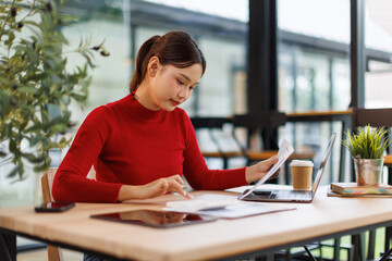 Smiling young asian woman sitting at desk working on laptop taking notes in notebook, happy...