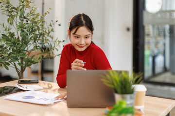 Smiling young asian woman sitting at desk working on laptop taking notes in notebook, happy...
