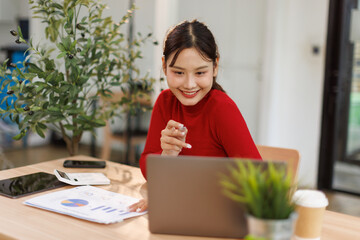Smiling young asian woman sitting at desk working on laptop taking notes in notebook, happy...