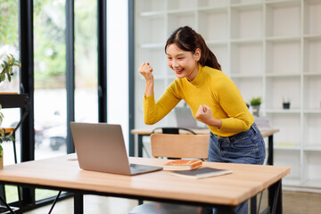 Obraz premium Happy young Asian business woman celebrating success at work in office looking at laptop excited about financial trading growth, goals achievement good online results, screaming yes watching game. 