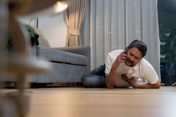 Asian man having chest pain and calling emergency on phone while lying on the floor at home, heart...