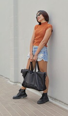 Asian woman exudes confidence as she leans against a wall, striking a casual yet stylish pose with a modern black tote bag hanging from her hands