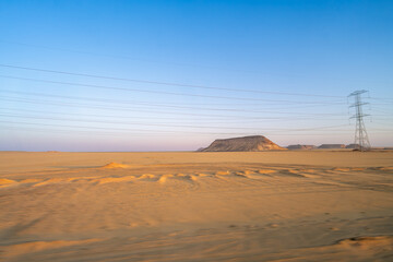 The vast Aswan desert landscape at dawn, showcasing unique rock formations and a prominent power line tower under a clear sky, along the route from Aswan to Abu Simbel.