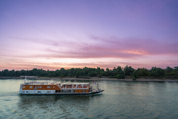 Obraz premium A peaceful evening on the Nile River, Egypt, with a cruise boat sailing towards Esna Lock. The sky displays soft hues of purple and pink as the sun sets over the lush green banks.