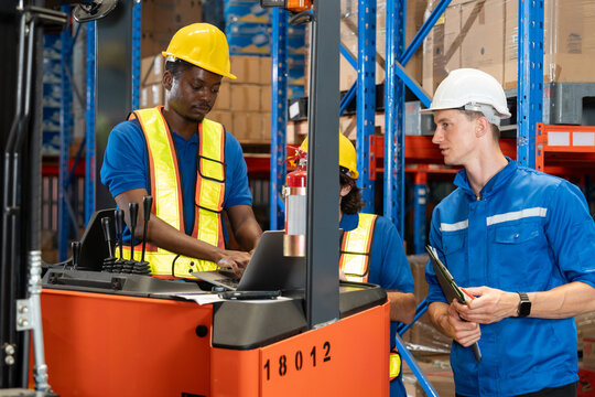 Group of warehouse workers including African male forklift operator, Caucasian male supervisor, and technician in safety helmets and vests using laptop and discussing logistics tasks in storage area.