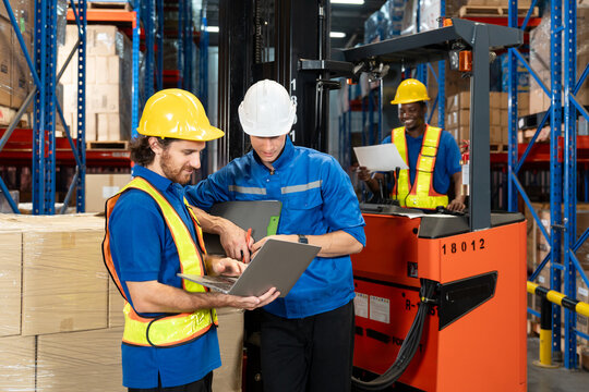 Three male warehouse workers in safety helmets and reflective vests discussing supply chain data using laptop and clipboard while forklift operator in background checking document in storage area.