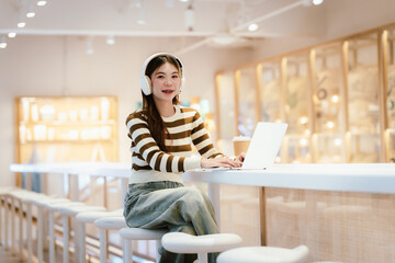Young Asian woman sitting in a cozy café, smiling while using her smartphone and laptop, enjoying a hot drink, studying, working, and relaxing in a modern setting.
