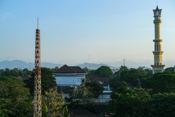 Obraz premium Cityscape view featuring a tall minaret and densely packed rooftops beneath a hazy sky with mountains in the distance, highlighting the urban texture of Mataram city in Lombok.