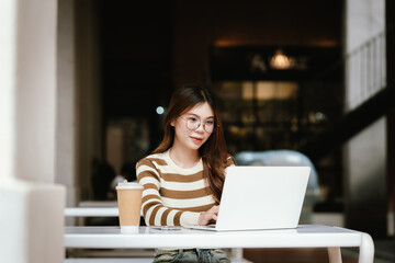 A young woman with glasses and a coffee cup uses her smartphone at a table, likely performing an...