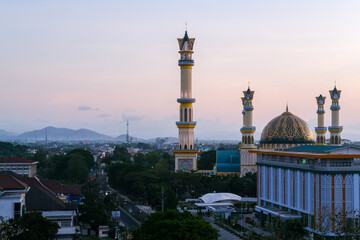 Fototapeta premium Golden twilight blankets the majestic mosque in Mataram city, Lombok, highlighting elegant minarets, vibrant streets, and surrounding hills under a serene pastel sky. Masjid Islamic Center Mataram.