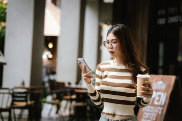 A stylish young woman, possibly an influencer, holds a coffee cup and uses her smartphone outdoors in an urban cafe environment.