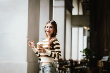 A stylish young woman, possibly an influencer, holds a coffee cup and uses her smartphone outdoors...