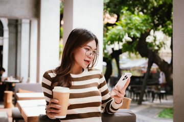 A stylish young woman, possibly an influencer, holds a coffee cup and uses her smartphone outdoors in an urban cafe environment.