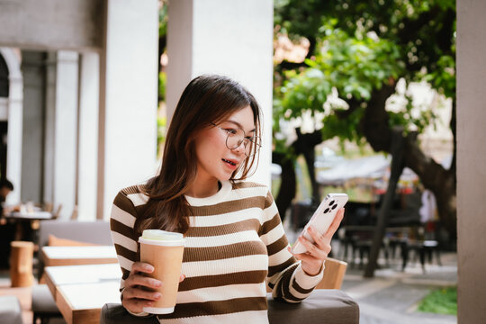 A stylish young woman, possibly an influencer, holds a coffee cup and uses her smartphone outdoors in an urban cafe environment.