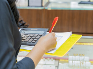 A woman is writing with a red pen on a financial sheet while using a calculator, standing in a jewelry store. Concept of price checking, retail inventory, business calculation, and data entry.