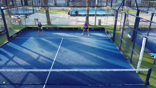 Two women playing padel on a blue outdoor court during sunny day. Energetic match in modern sports facility with glass walls and palm trees in the background.