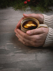 Christmas vibes. Female hands in a white sweater hold a coffee mug with golden bokeh n on a wooden table, vertical image.